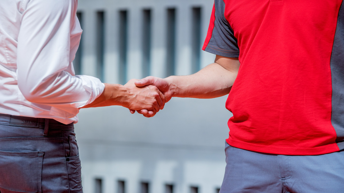 Zwei Personen schütteln sich die Hände, eine in einem weißen Hemd und eine in einem rot-grauen Hemd, die im Freien stehen.