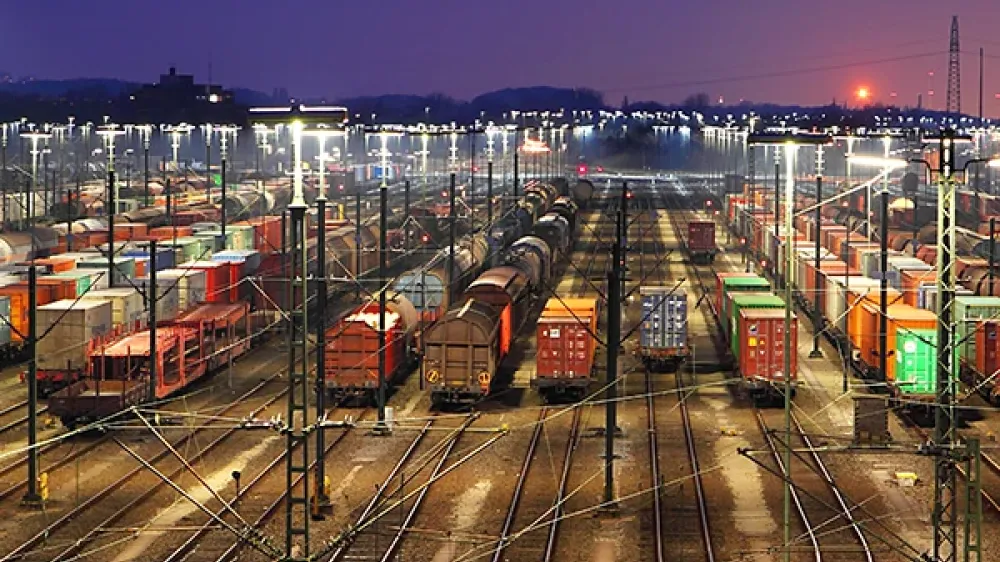 Recinto de una estación de ferrocarril con muchas vías, agujas y líneas eléctricas frente a la silueta de una ciudad.
