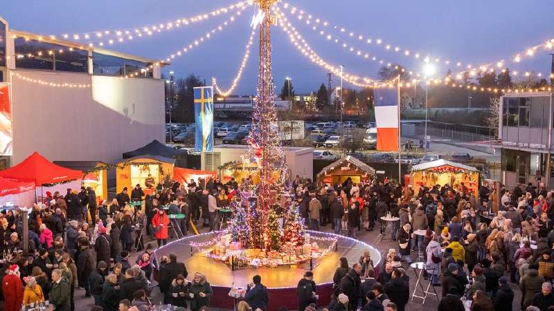 Una escena festiva de un mercadillo navideño muestra un árbol alto e iluminado, adornado con luces y adornos. Multitudes de personas vestidas con ropa de invierno se reúnen alrededor de los puestos festivos, disfrutando del ambiente animado y alegre. Guir