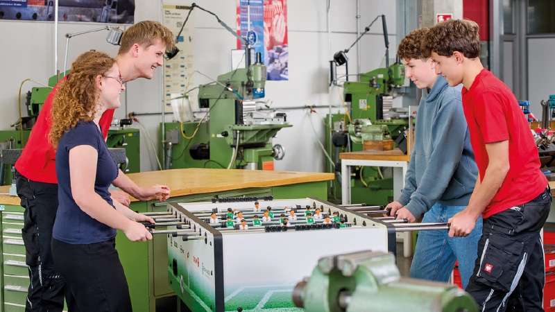 Cuatro jóvenes, dos a cada lado, juegan al futbolín en un taller industrial. Están sonrientes y concentrados, con maquinaria verde al fondo.