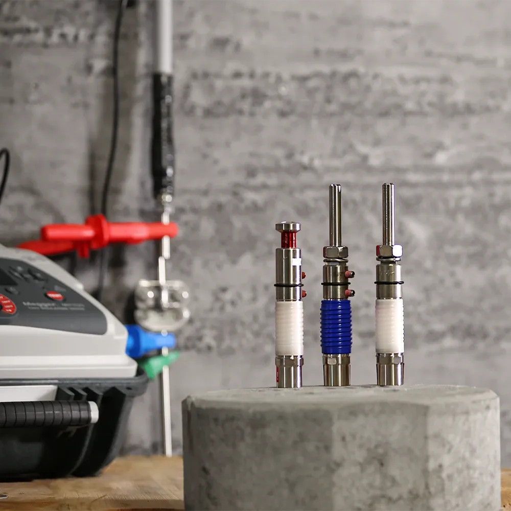 Tres objetos cilíndricos de metal con componentes rojos, azules y blancos sobre una superficie de hormigón delante de un equipo con un panel de control.