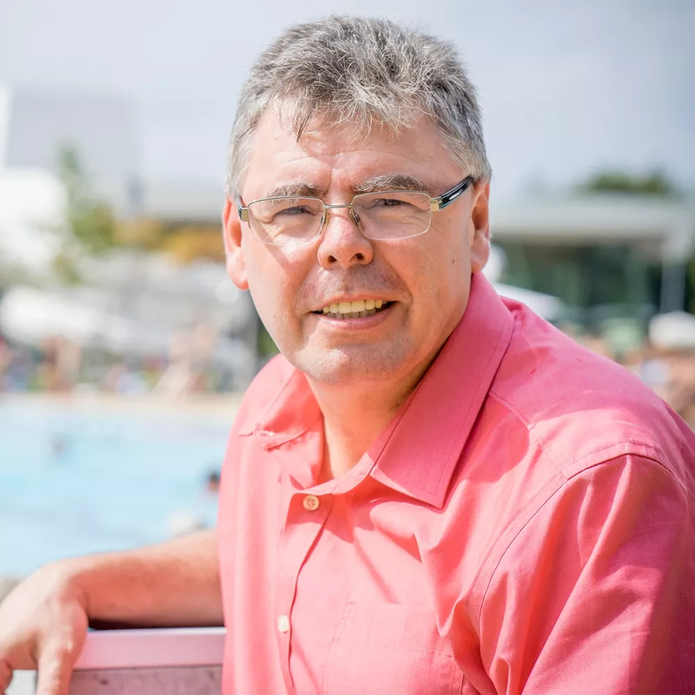 Portrait of a man wearing glasses and a pink shirt, leaning on a railing near an outdoor pool in daylight.