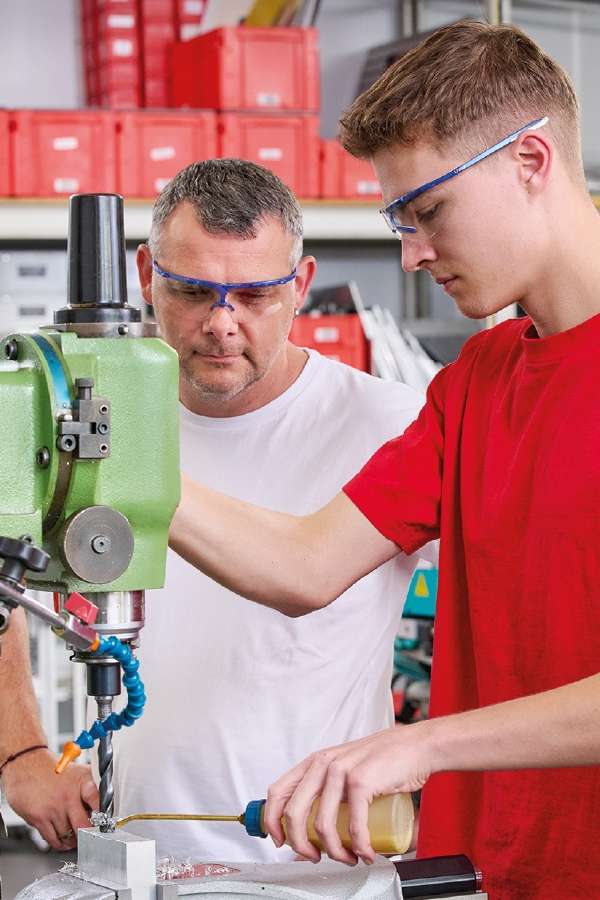 Dos hombres que llevan gafas de seguridad trabajan con un taladro de columna verde en un taller. Uno maneja la máquina, mientras que el otro supervisa, con la mirada fija en ella.