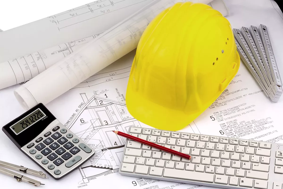 Yellow safety helmet on architectural blueprints with calculator, keyboard, ruler, and pencil, representing technical planning and construction work.