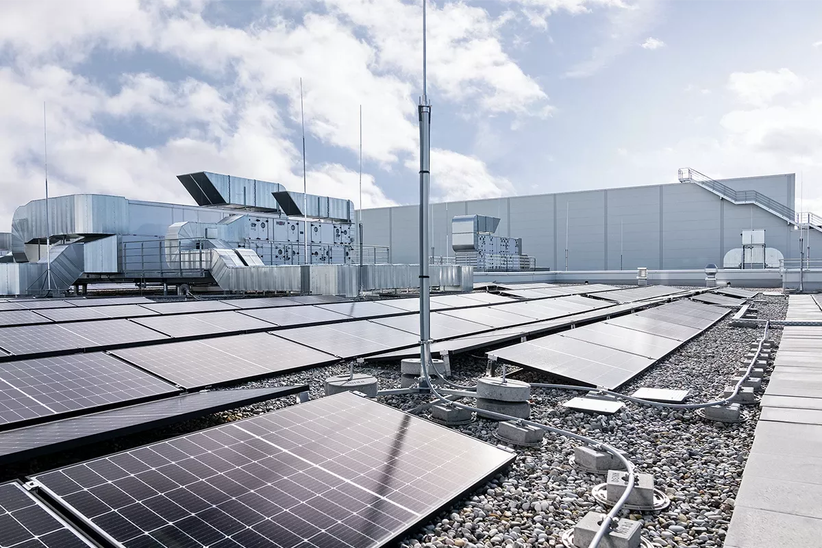 Rooftop photovoltaic system with solar panels, lightning protection mast, and cable routing on a commercial building roof under a cloudy sky.